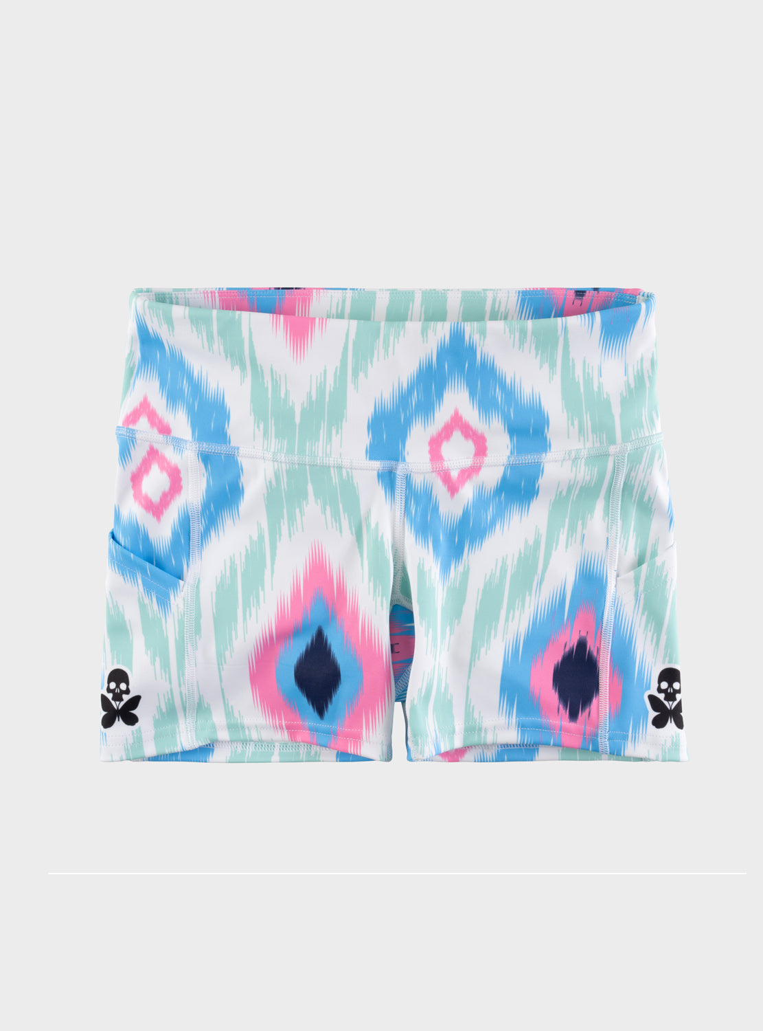 A pair of women's athletic shorts featuring a colorful ikat pattern in pink, blue, and mint green on a white base, with small black skull-and-butterfly logos on the lower sides.