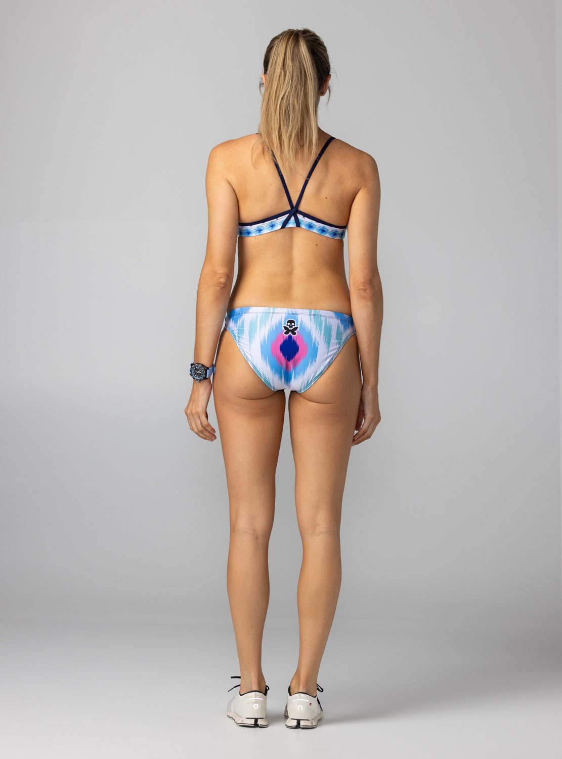 A woman shown from the back wearing a bikini. The bikini bottom has a blue, white, and pink tie-dye pattern with a central pink and blue diamond design and a small skull-and-butterfly logo.
