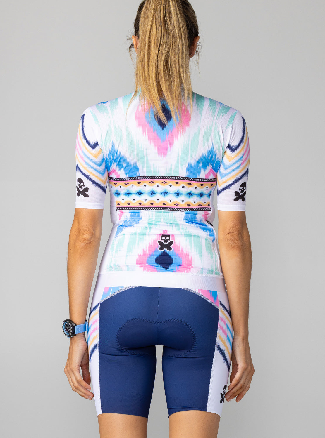 A woman shown from the back wearing a short-sleeve cycling jersey and matching cycling shorts. The jersey has a white base with a colorful geometric ikat-style pattern in pink, blue, orange, and yellow, along with small black skull-and-butterfly graphics on the sleeves.