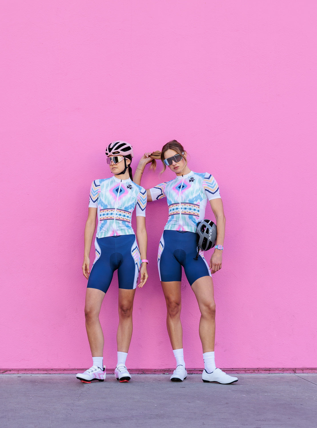 Two cyclists posing in matching light blue and white patterned cycling jerseys with navy blue shorts, standing against a solid pink wall.