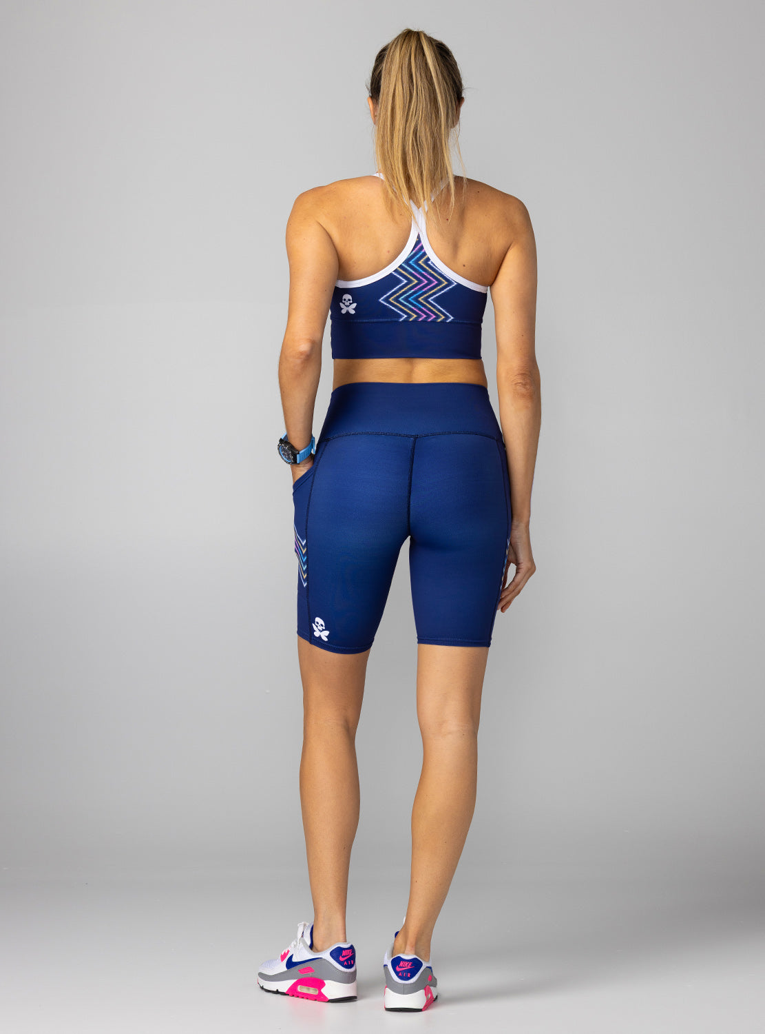 A woman shown from the back wearing a matching athletic set consisting of a navy blue and white patterned sports bra and navy blue biker shorts, paired with white, blue, and pink sneakers, against a solid gray studio background.
