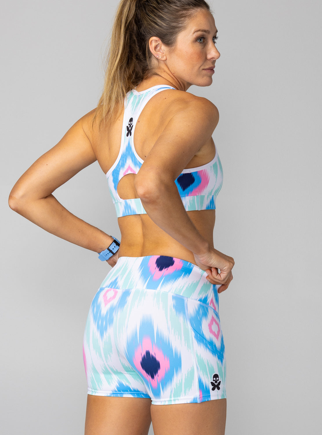A woman wearing a matching athletic set with a sports bra and shorts featuring a blue, pink, and white ikat pattern, shown from a three-quarters back view against a solid gray background.