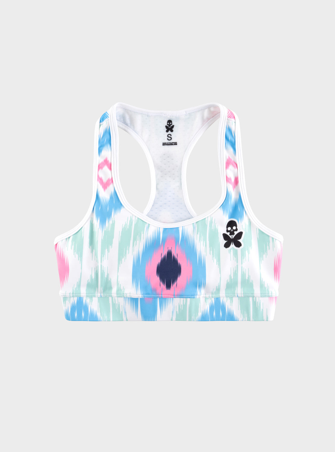 A women's racerback sports bra with a white base and a pastel ikat pattern in pink, light blue, and mint green. It features a small black skull-and-butterfly logo on the left chest and a black brand label on the inside neck.