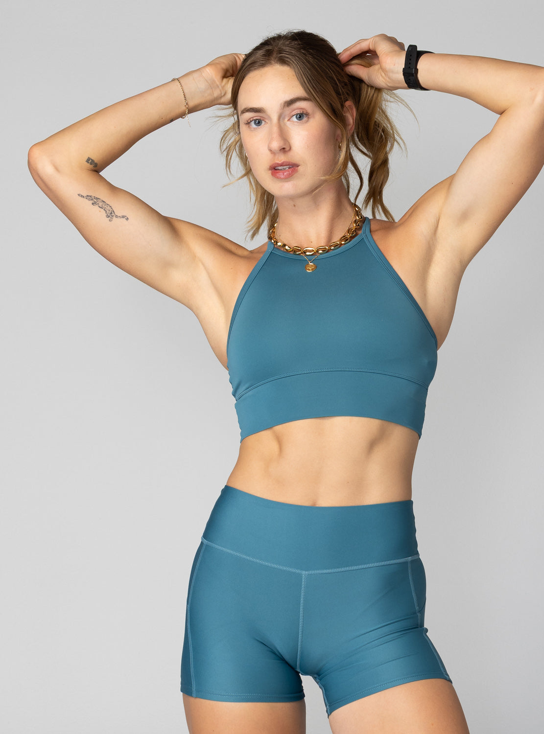 A woman modeling a matching teal blue athletic crop top and high-waisted shorts set against a solid gray studio background.