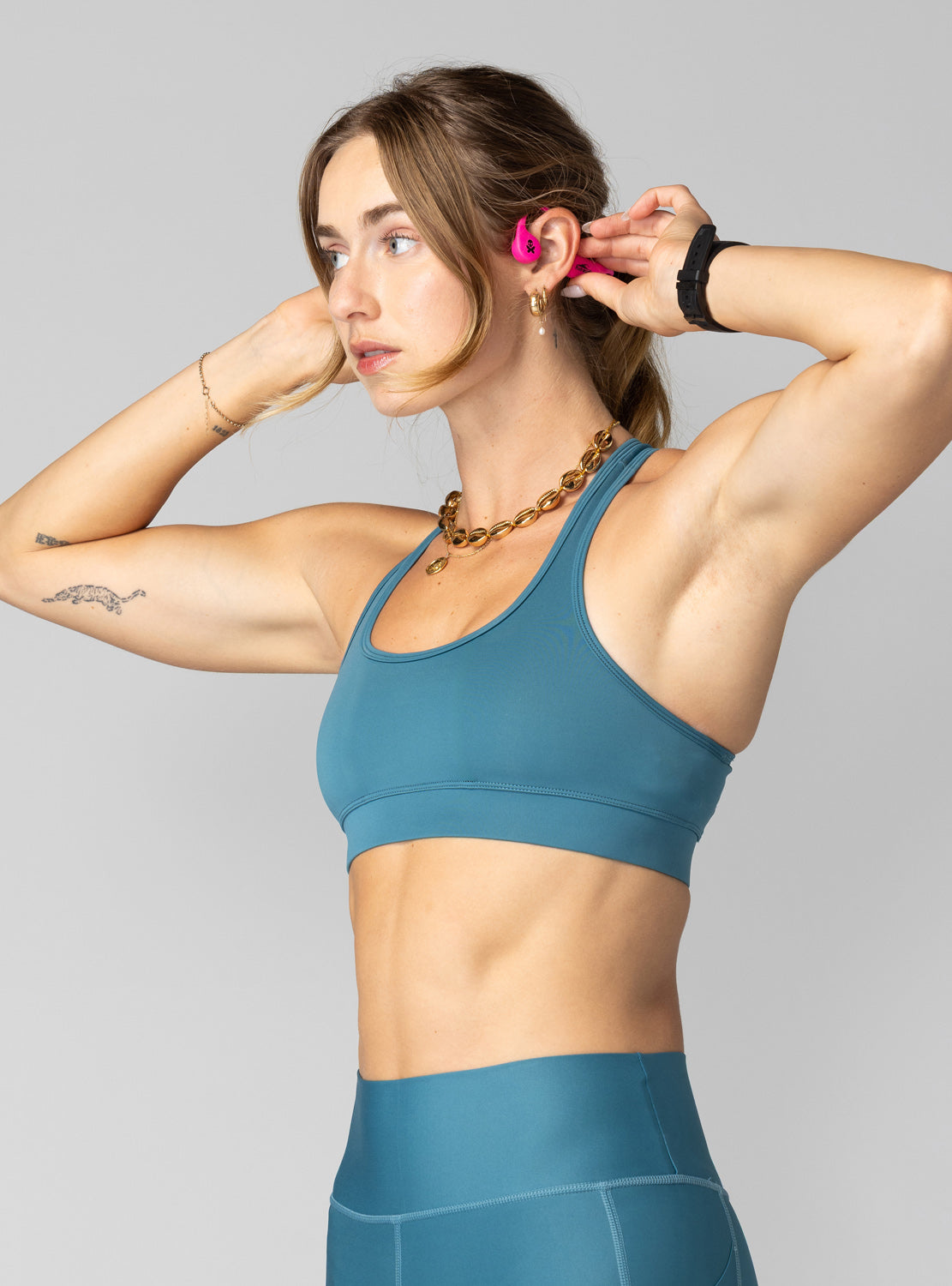A woman wearing a teal blue sports bra and matching high-waisted leggings, posing against a solid gray background.