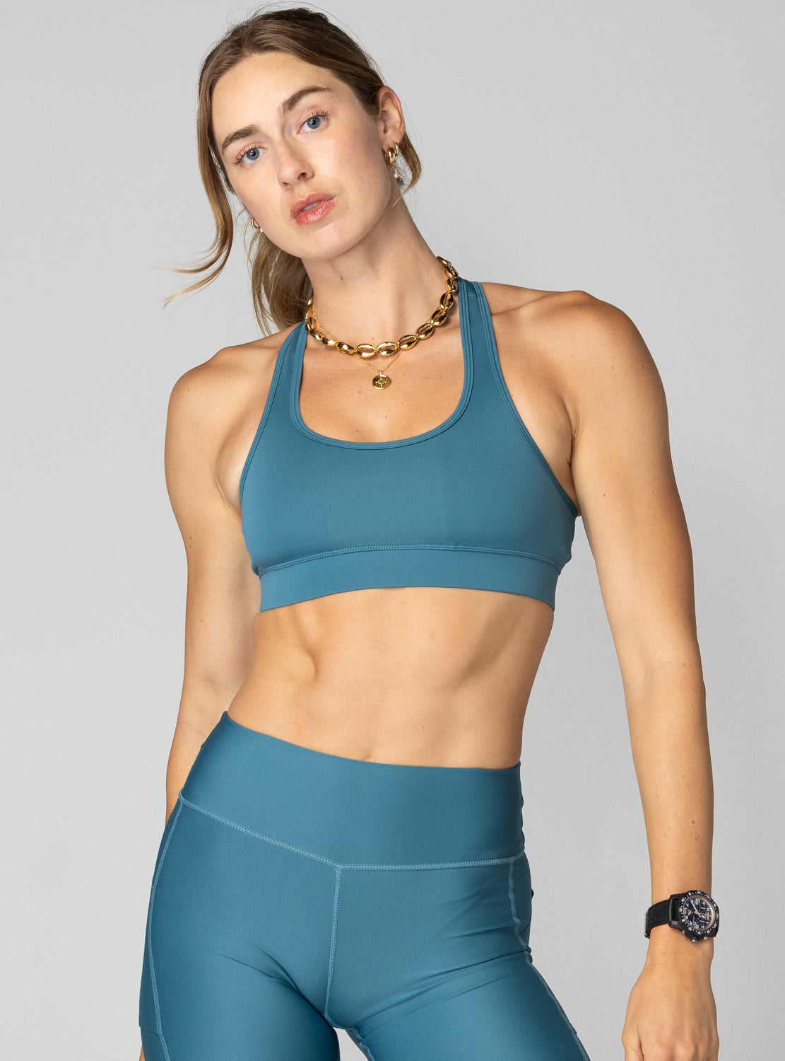 A woman modeling a teal blue athletic sports bra and matching high-waisted leggings against a solid gray studio background.
