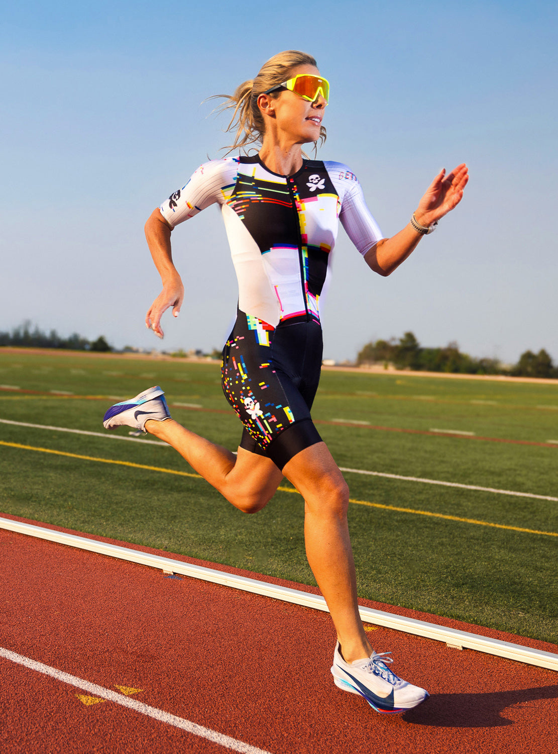 A woman running on an athletic track wearing a white, black, and multicolor cycling jersey, matching black shorts with colorful accents, yellow sunglasses, and white running shoes on a sunny day.