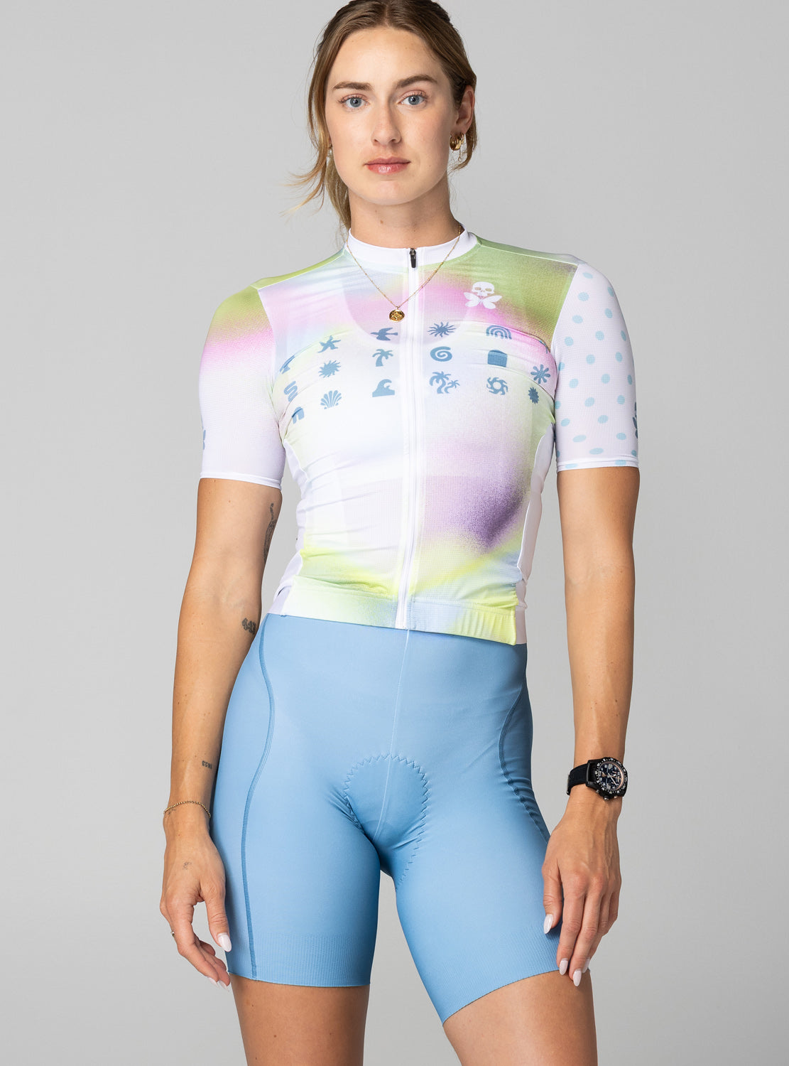 A woman modeling a short-sleeve cycling jersey and matching light blue cycling shorts against a solid gray studio background. The jersey features a pastel multicolor gradient with blue graphic icons across the chest, and the shorts are a solid light blue.